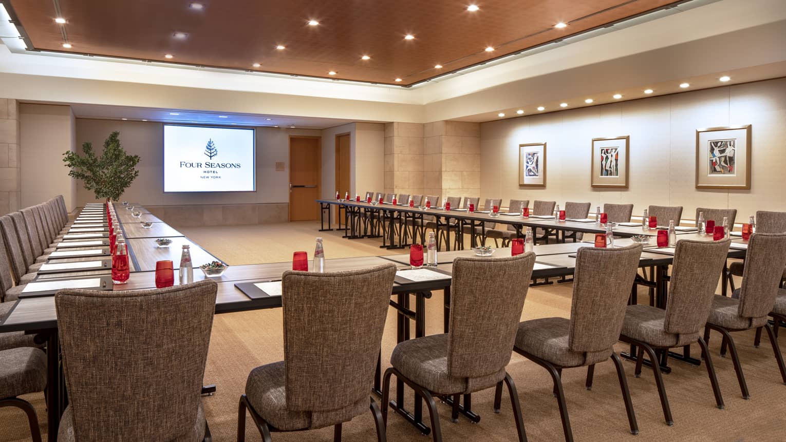 Multiple long, rectangular boardroom tables are placed together to form a "U" shape facing a presentation screen and are set with a black folder, white paper, pen and clear bottles filled with a red soda. 
