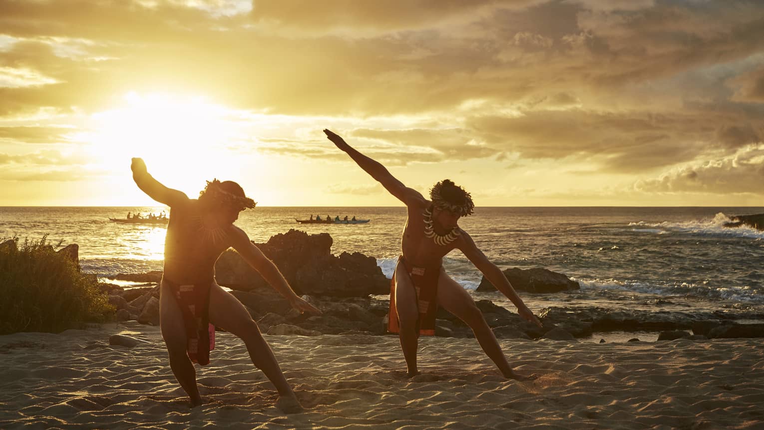 Two Hawaiian dancers with arms outstretched pose on sandy beach at sunset