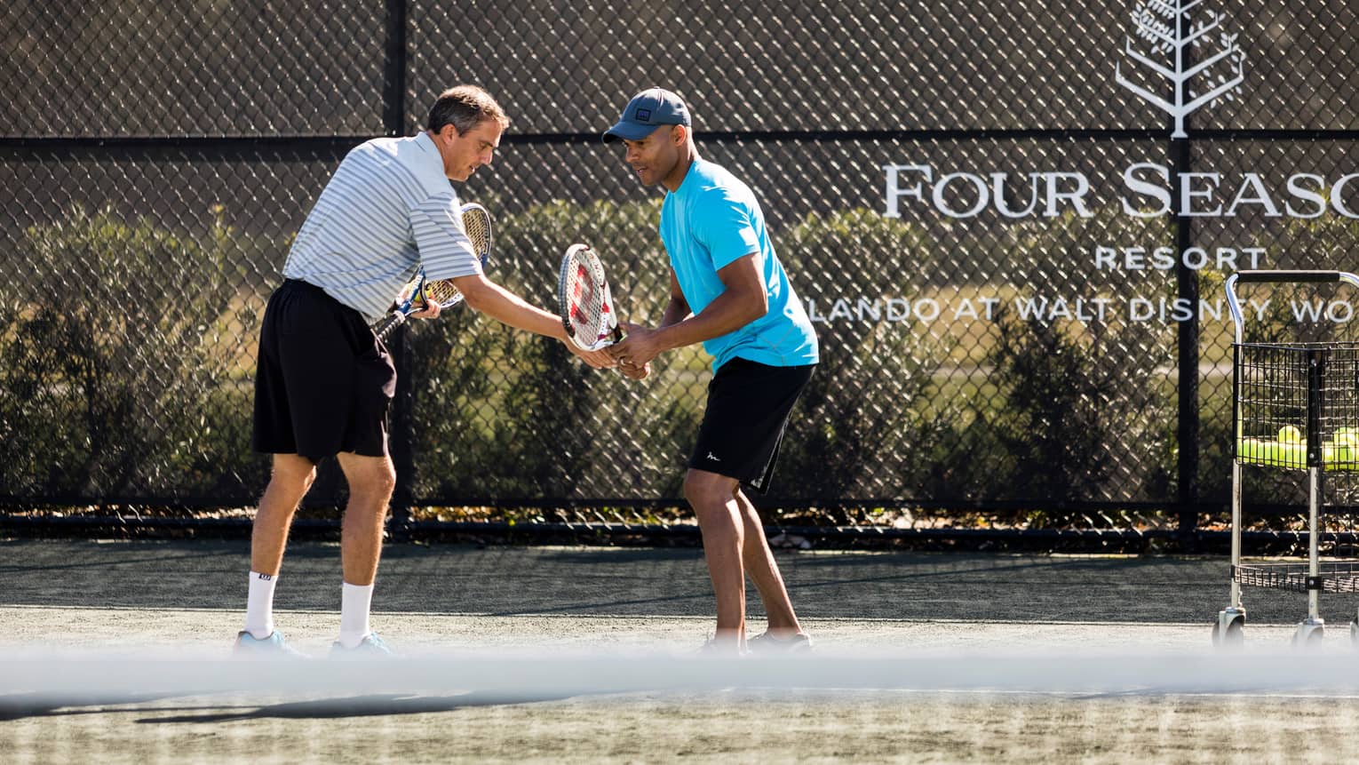 Tennis pro helps man adjust his racket on tennis court by Four Seasons Resort logo on fence