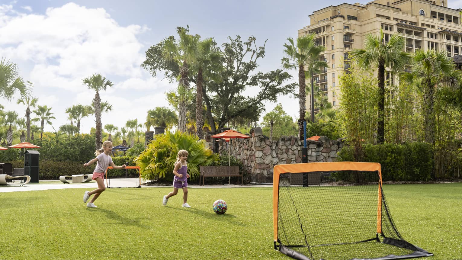 Two young girls playing soccer near palm trees.