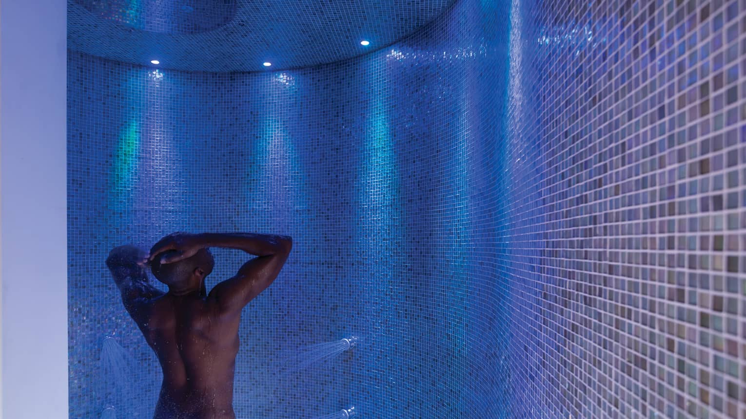 Man rinses off in tile spa shower under blue, purple lights