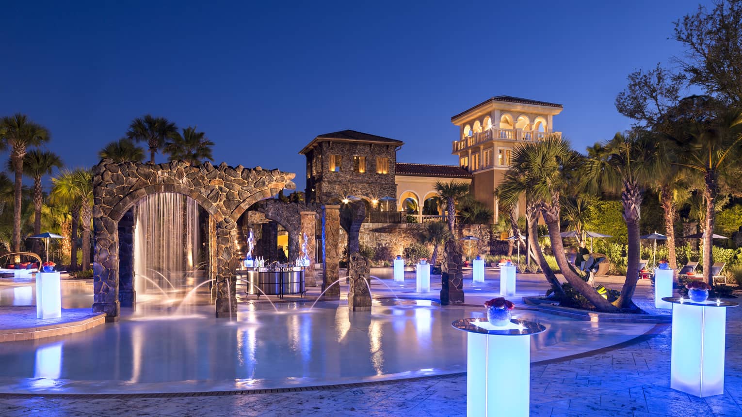 Blue glowing tables illuminate an outdoor bar area with palm trees and water fountains