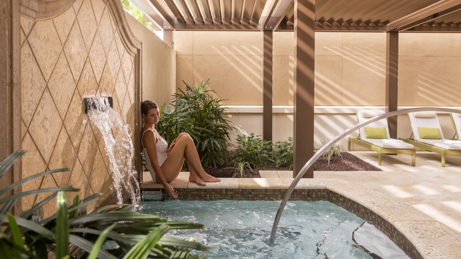 Woman wearing swimsuit leans against tile wall with fountain by small spa pool