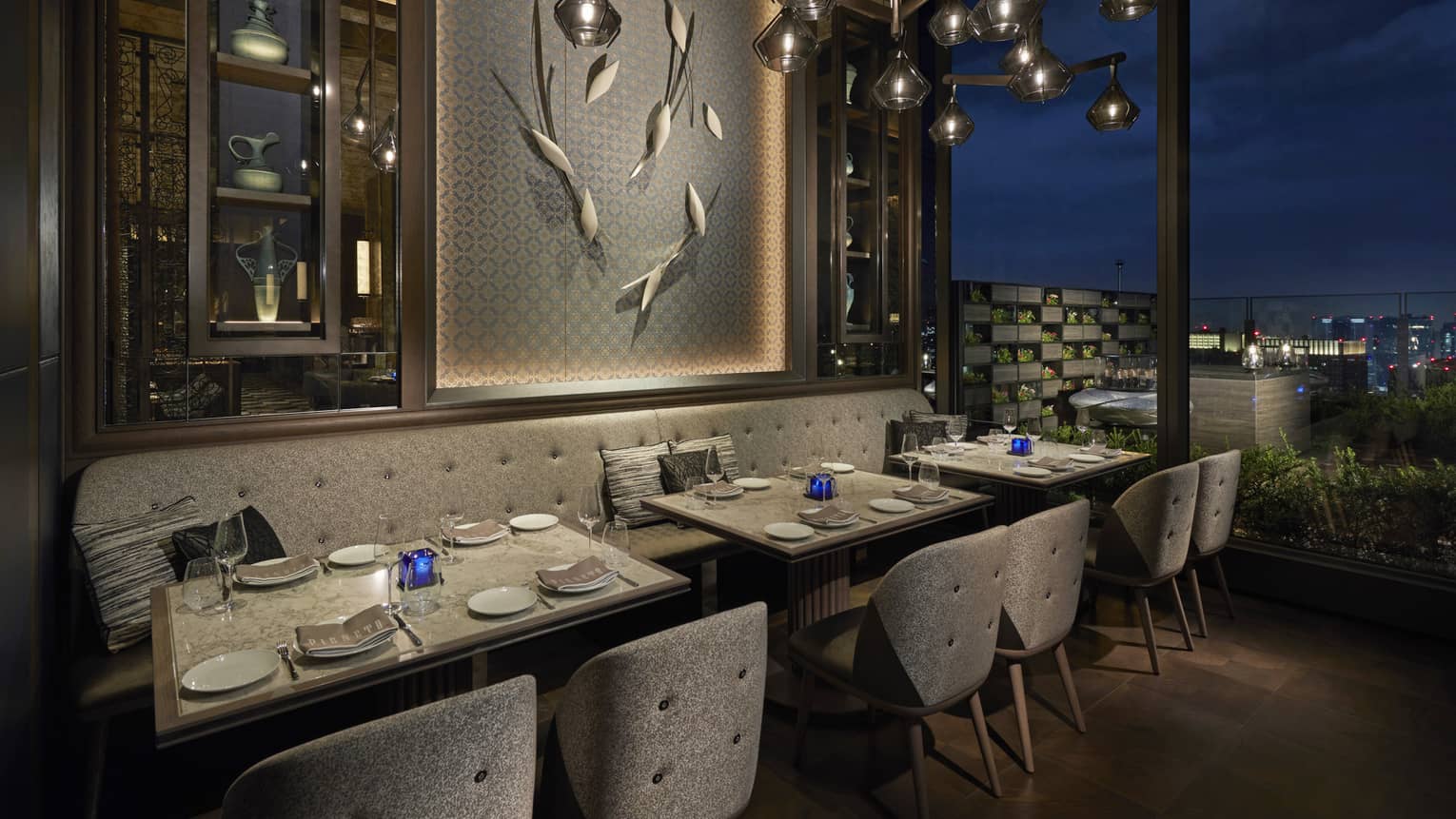 Tables set for two overlook nighttime skyline, floor-to-ceiling windows, intimate lighting, grey colour scheme 