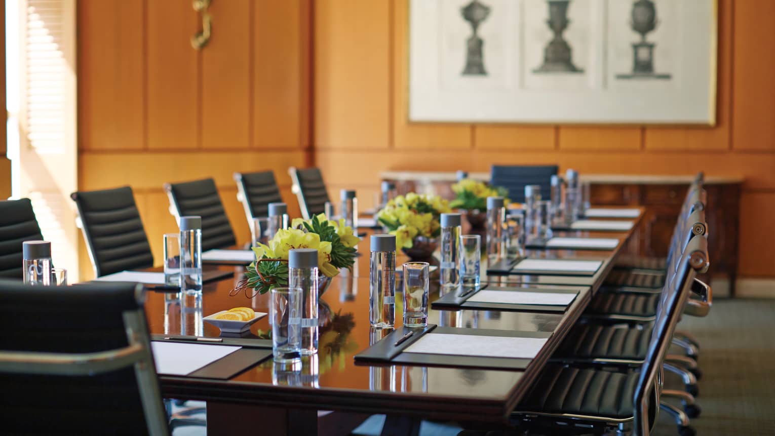 Large boardroom meeting table with yellow flowers, glassware under wood panel walls, art