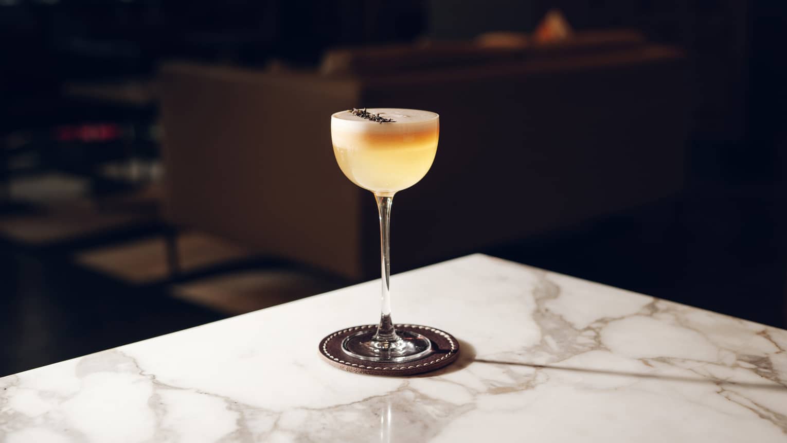Pale-yellow cocktail topped with foam and garnish in wine glass on marble table
