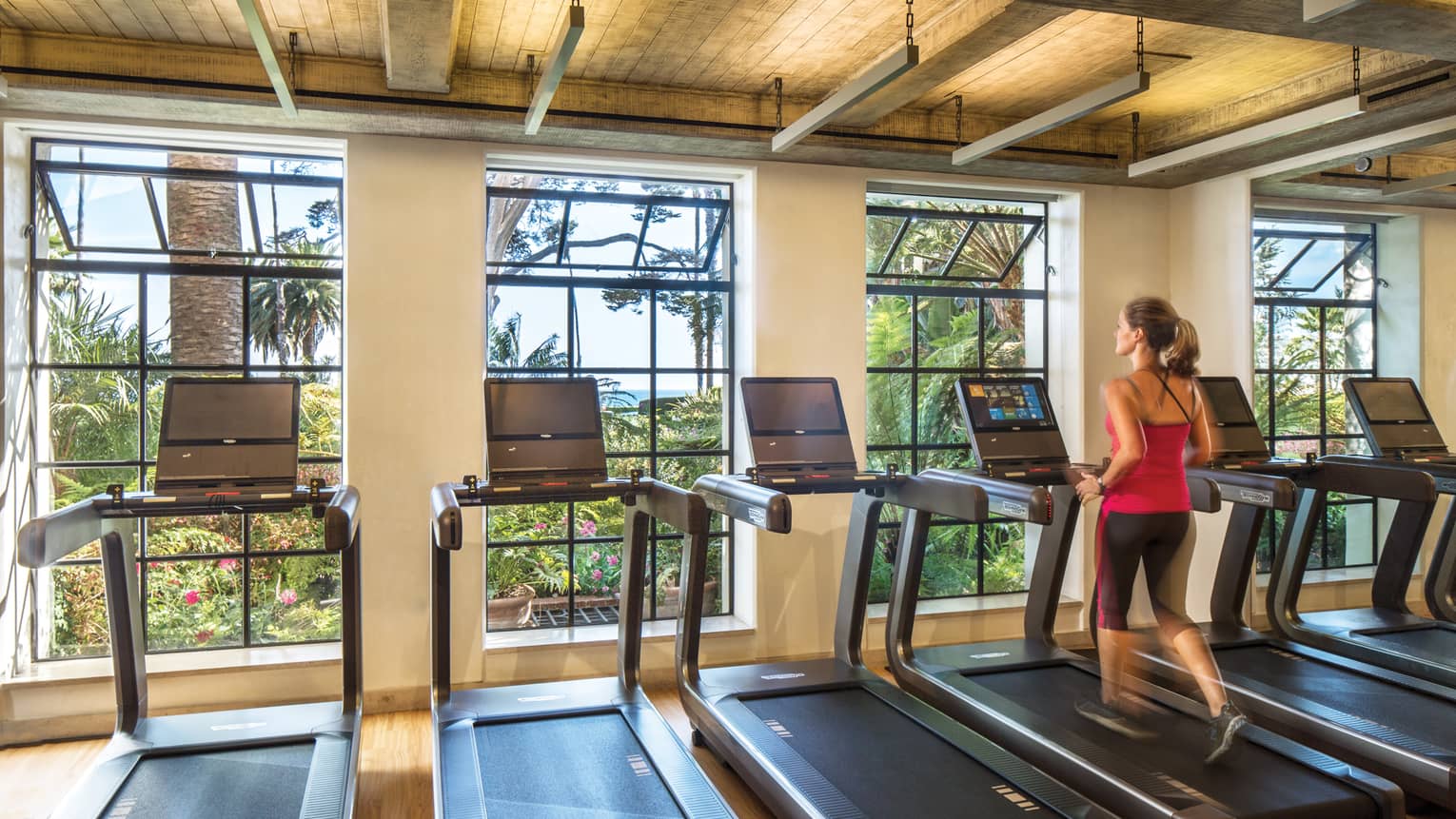Woman runs on row treadmill in row of machines by large open windows in Fitness Centre