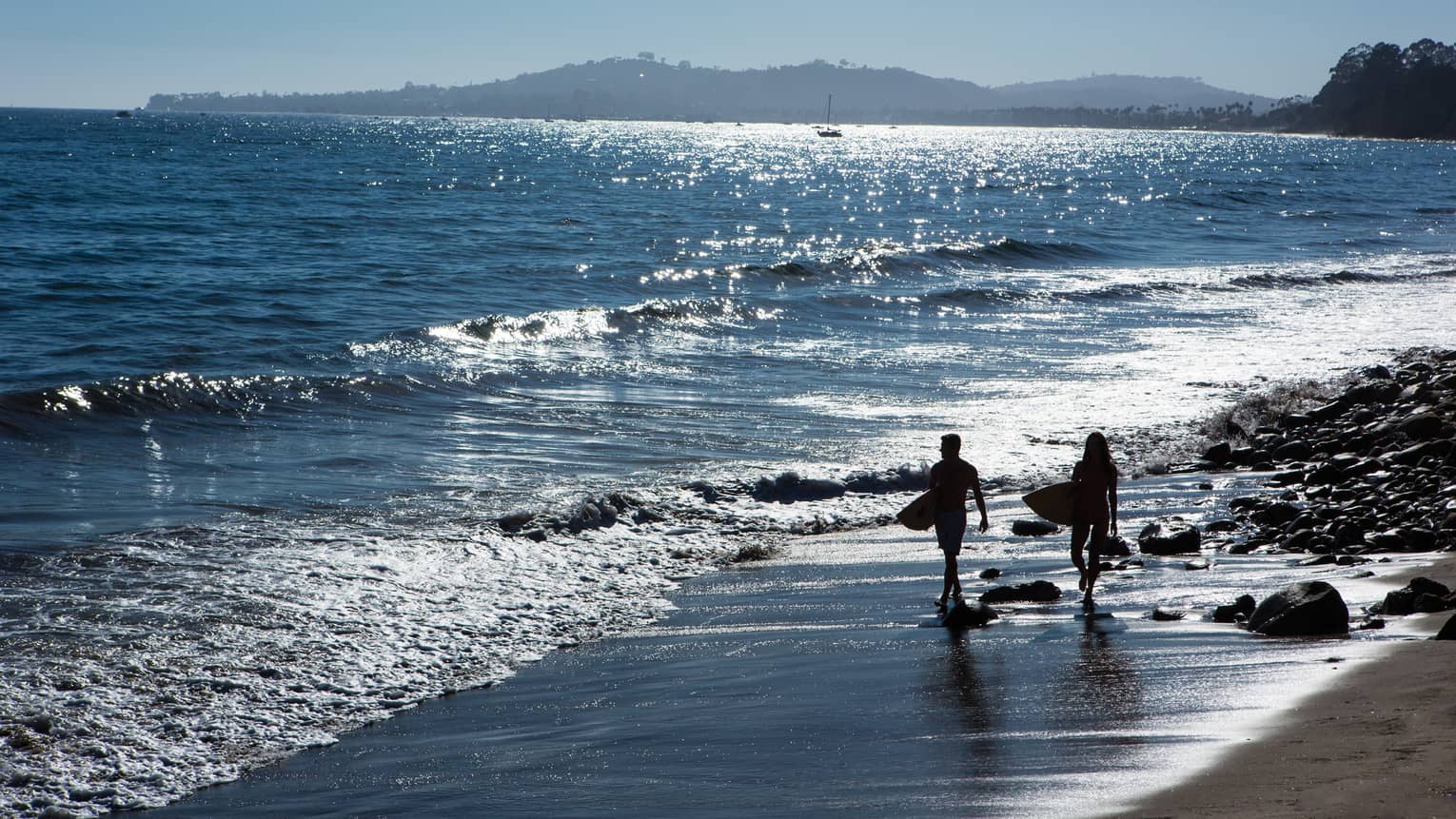 Silhouettes of two people carrying surfboards along beach shoreline
