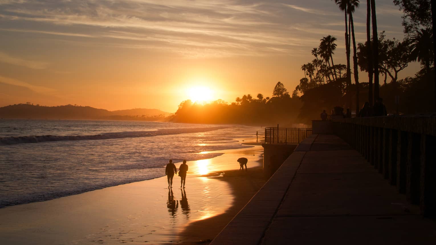 Silhouettes of three people walking along coast on sandy beach, orange sunset