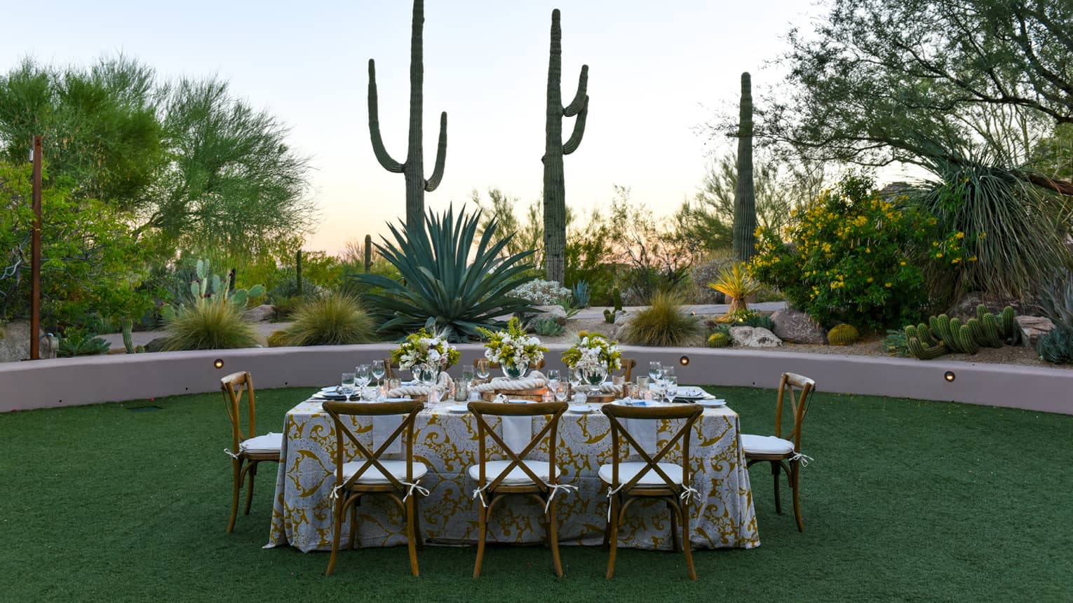 A rectangular table outside with a patterned cloth and wood chairs surrounded by cacti and trees.