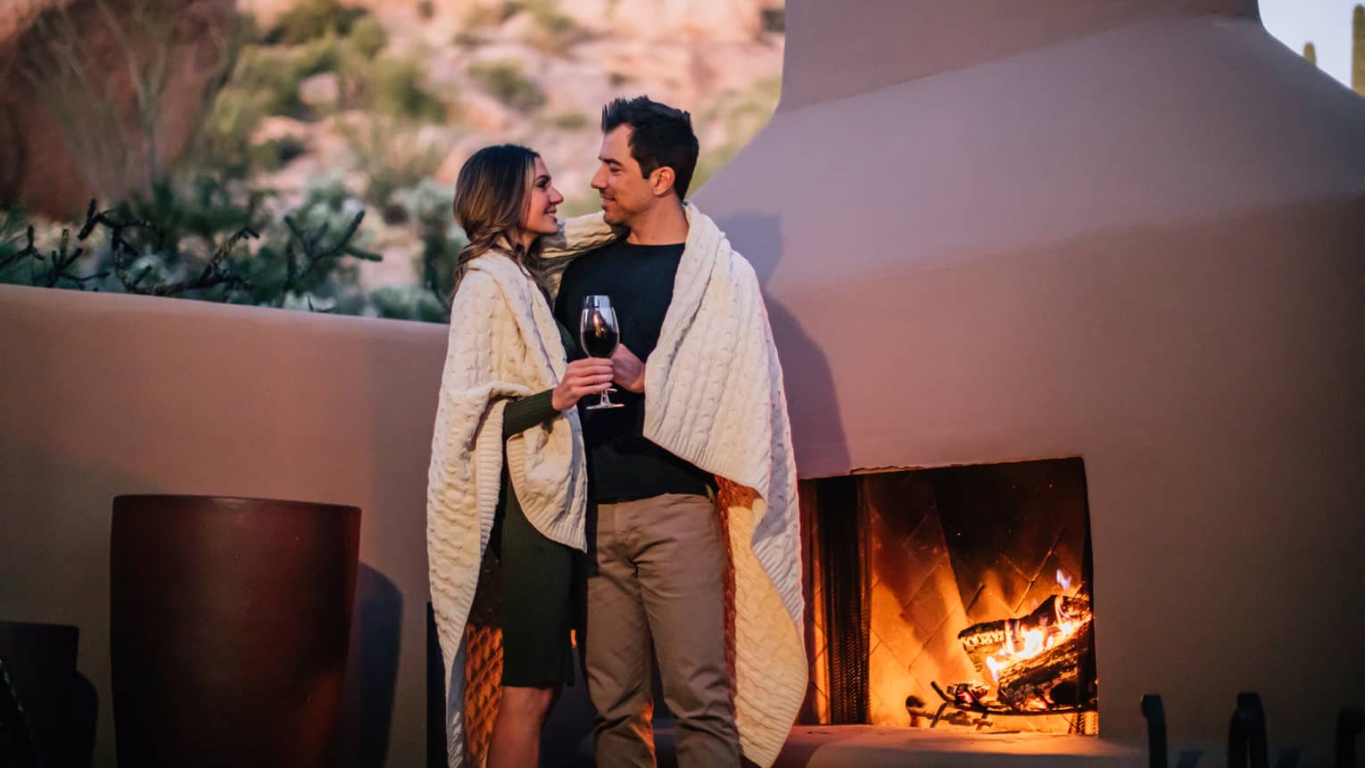 A woman and man with a blanket covering them next to a fire outside.