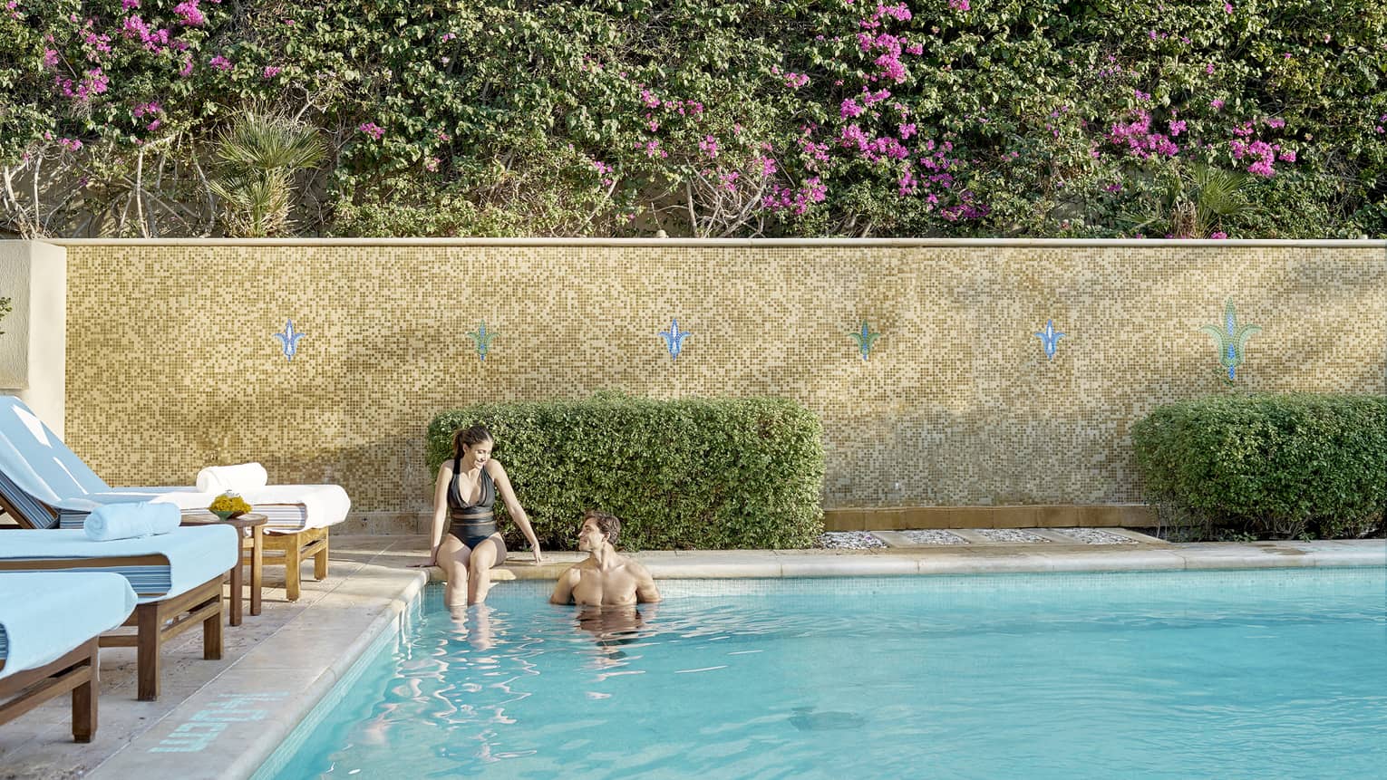 Couple relaxes poolside, row of lounge chairs and pink flowering hedges visible behind wall