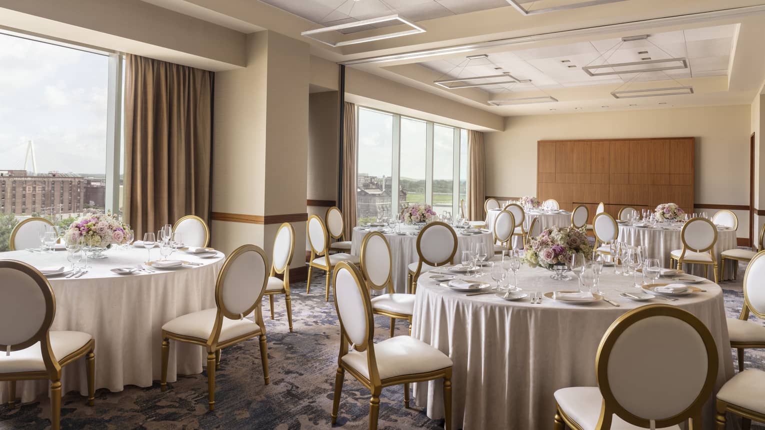 A room with round tables with table cloths, chairs, flower table settings, and large windows.