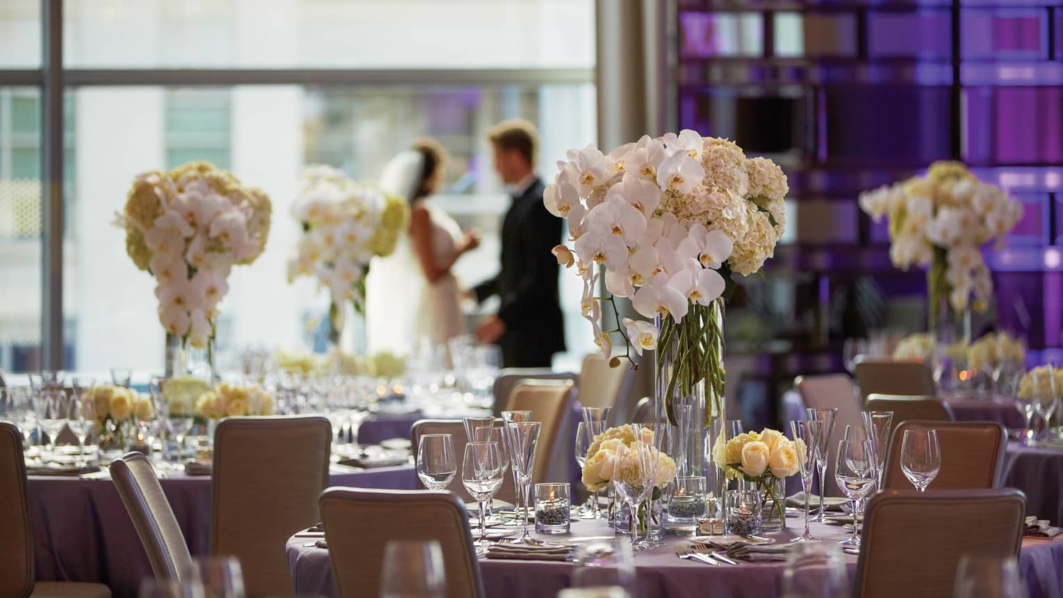 Wedding tables with white orchid centrepieces, bride and groom by sunny window in background