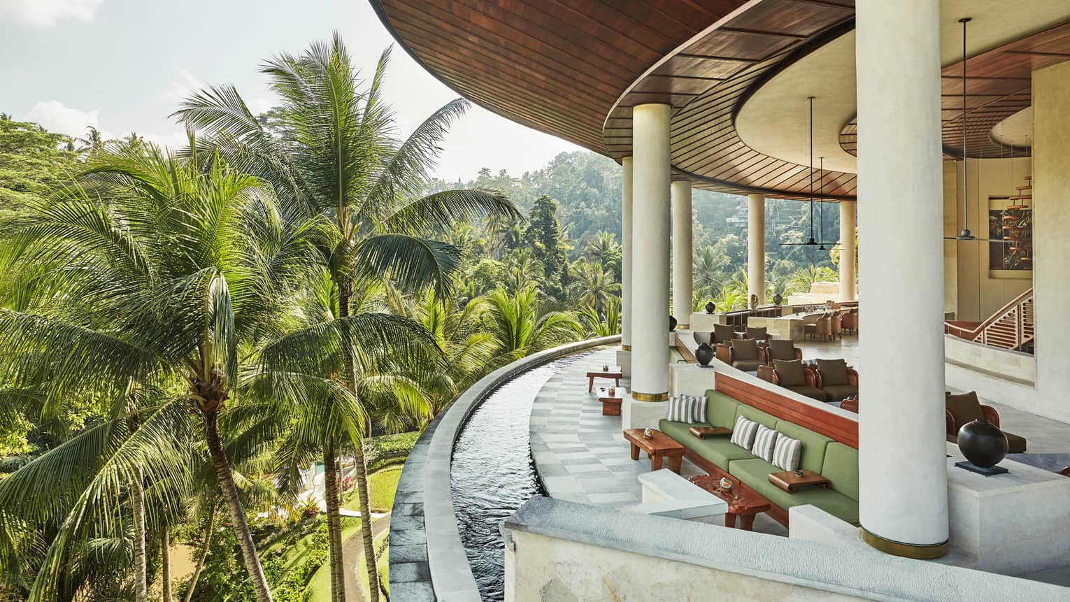 Curved outdoor balcony with water around ledge, green and wood sofas, chairs, vases, large palm leaves outdoors