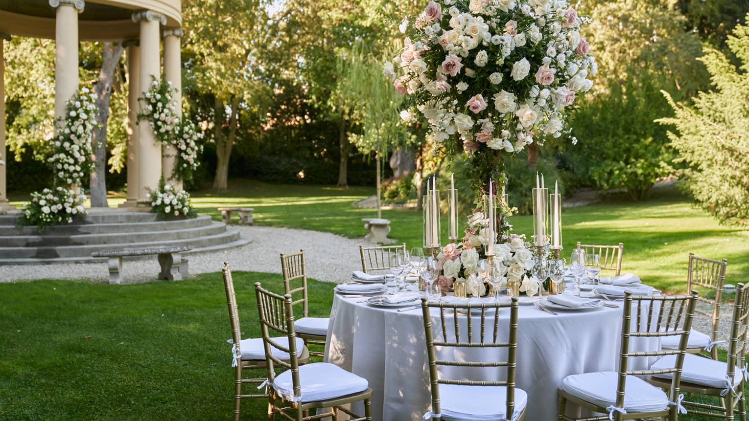 White formally set wedding reception table in garden with large white topiary and gazebo in backdrop
