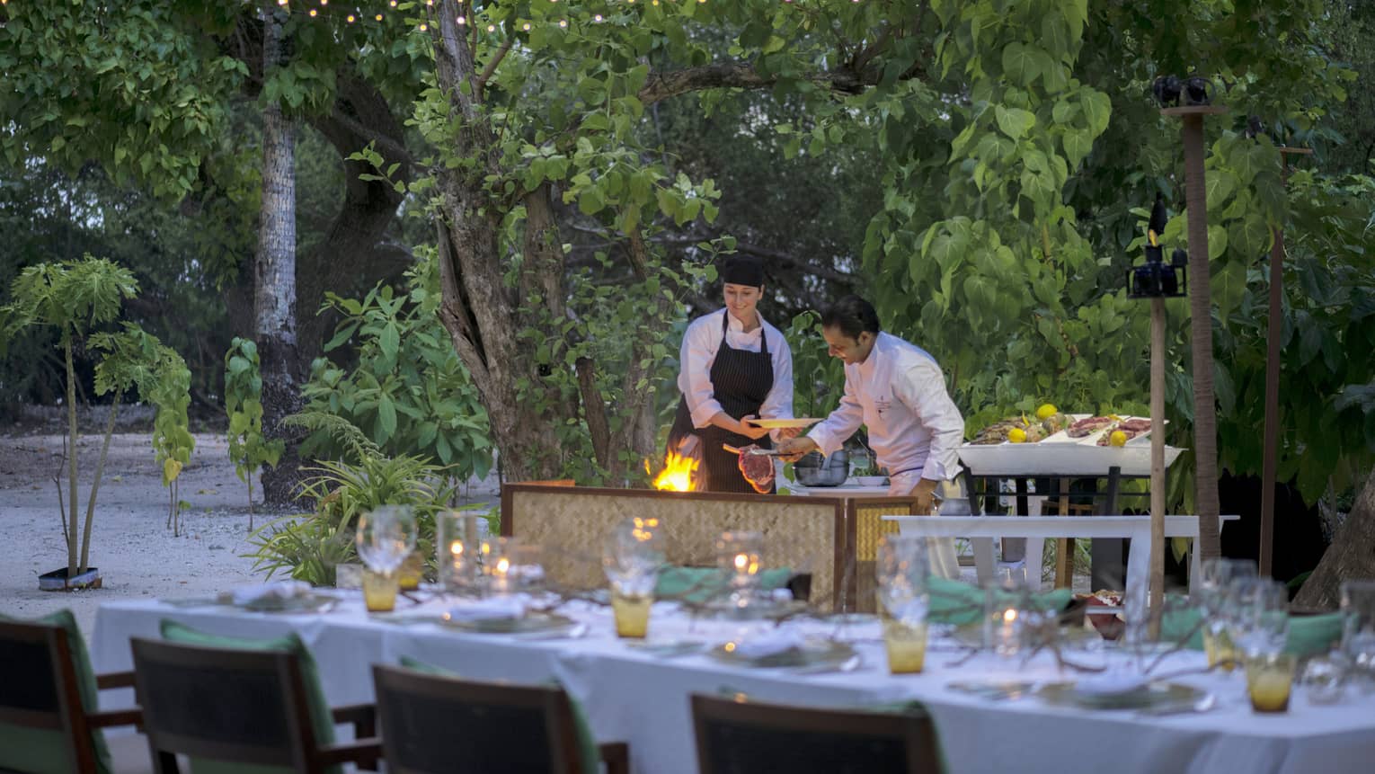 Chefs put large steak on flaming grill by long outdoor dining table, patio lights at night
