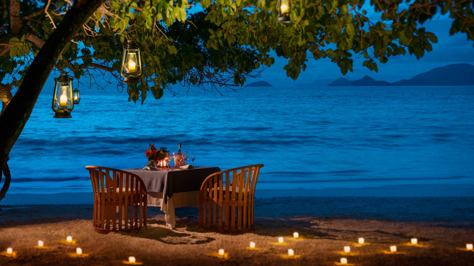 Round candle-lit dining table under tree, lanterns on beach at night