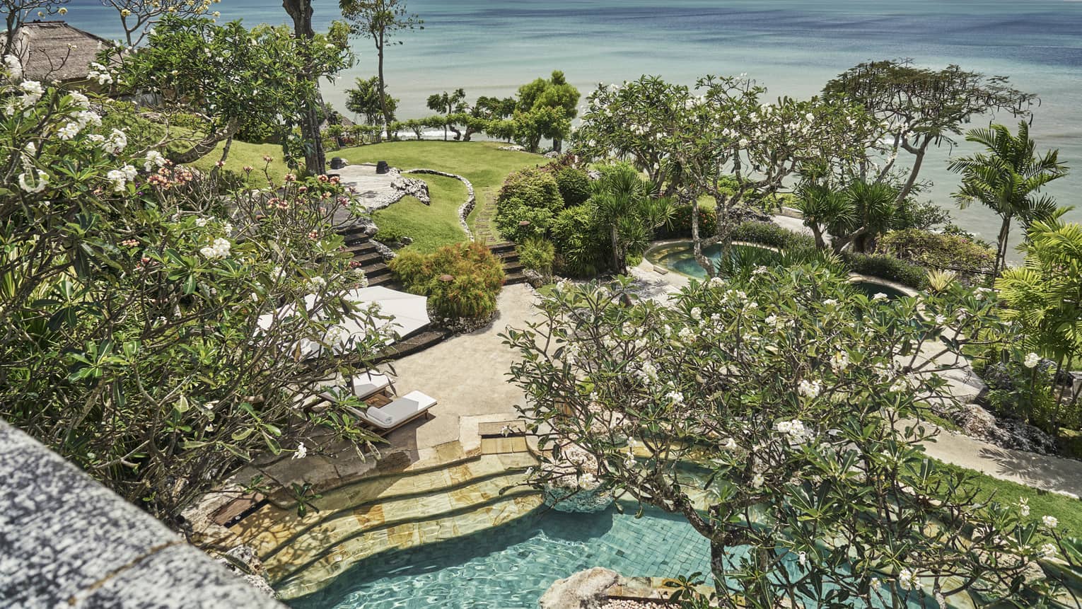 Aerial view through trees of resort stone patio and steps to swimming pool, green lawn, trees and blue ocean