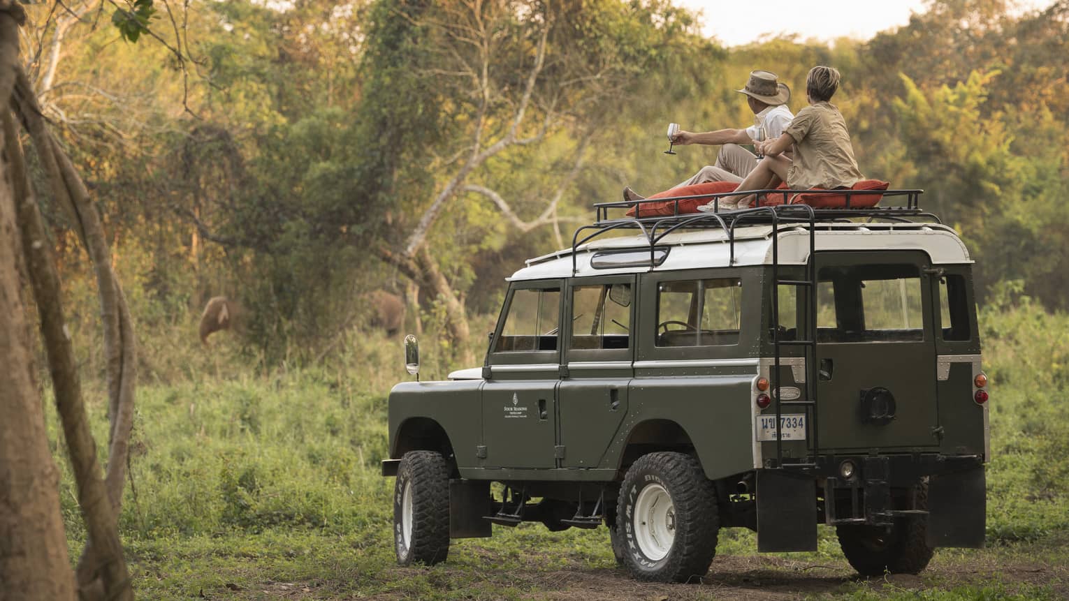 A couple sits on the roof of a Land Rover in the Bush, drinking a cocktail