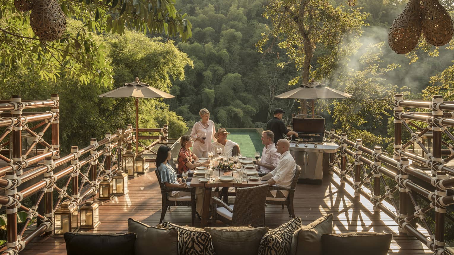 Smiling group of diners at patio table near chef at barbecue, surrounded by jungle