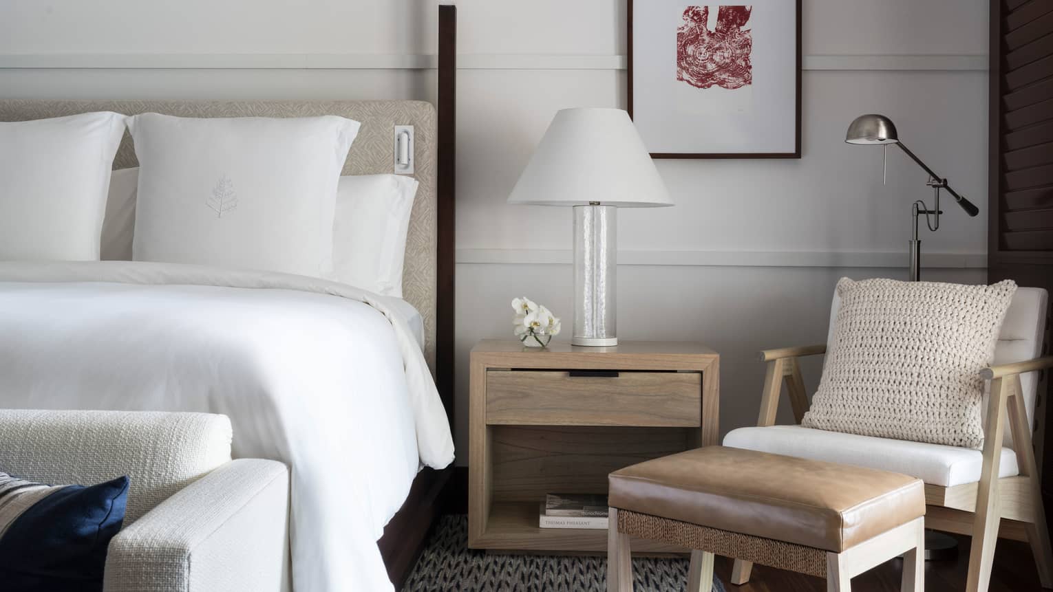 Close-up of white hotel bed, nightstand with white lamp, modern armchair and leather footstool