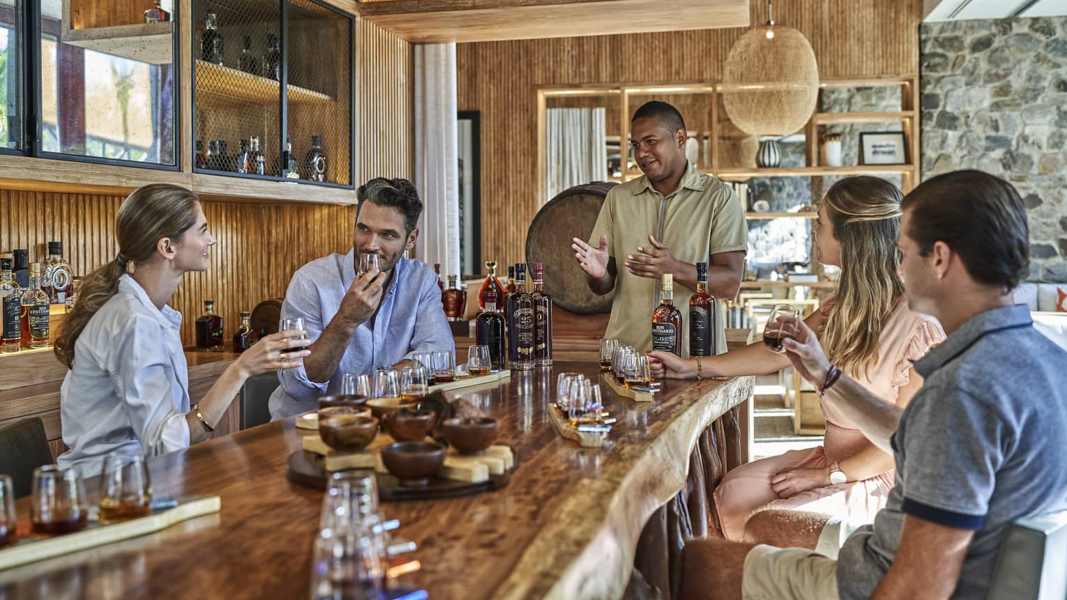 Four people sit around a live-edge wooden counter filled with selections of rum and chocolates as a rum master explains the flavors