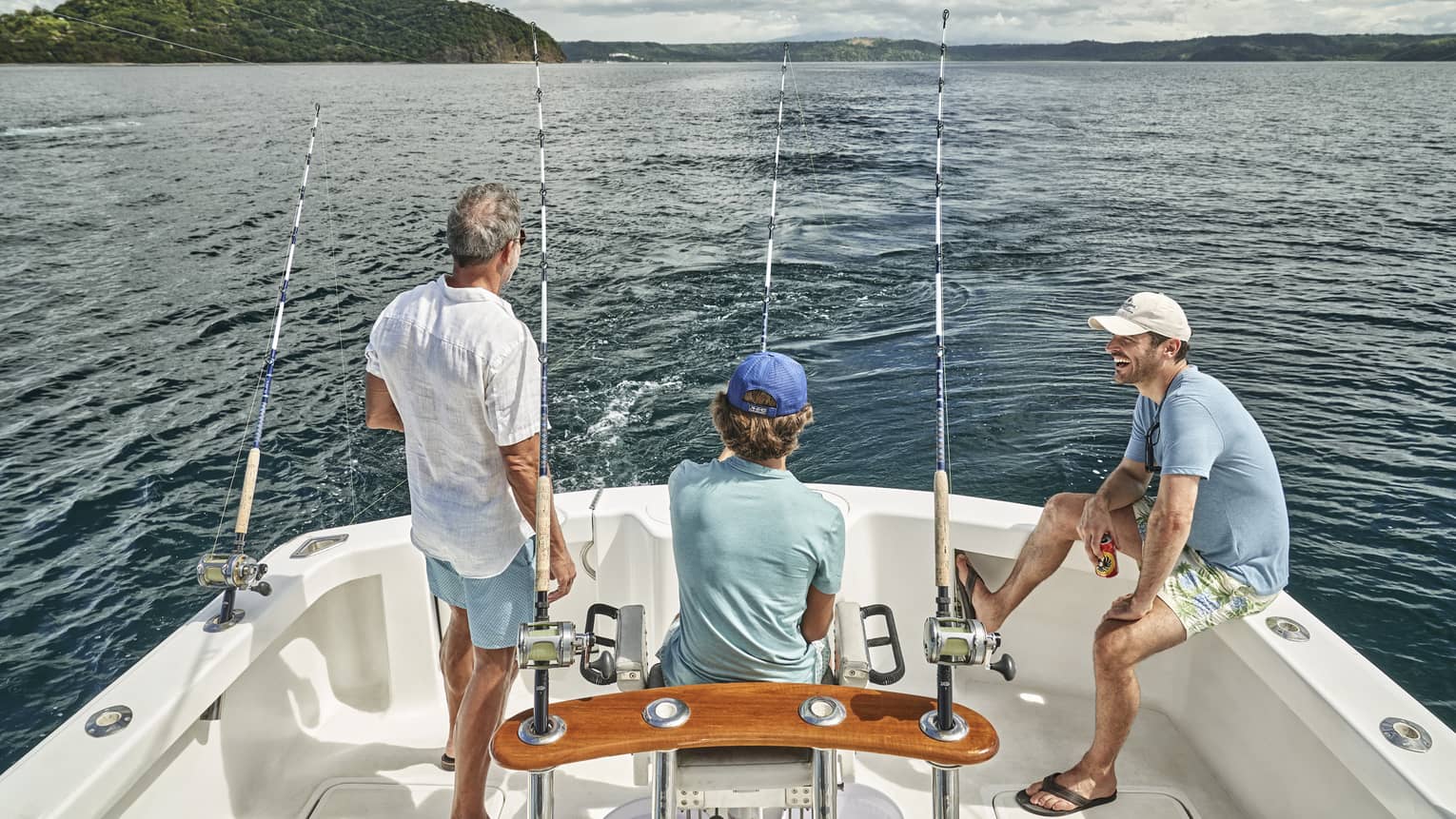 Three men of varying ages sit looking out at the water from the back of a boat outfitted with fishing rods