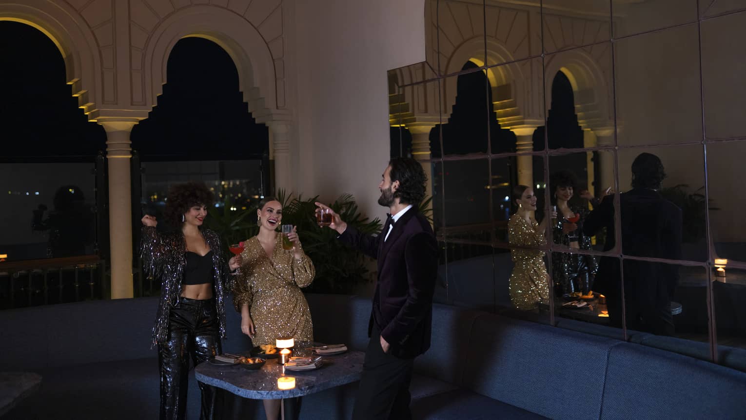 Two women and one man all dressed in cocktail attire raise a toast around a candlelit table next to a stone archway