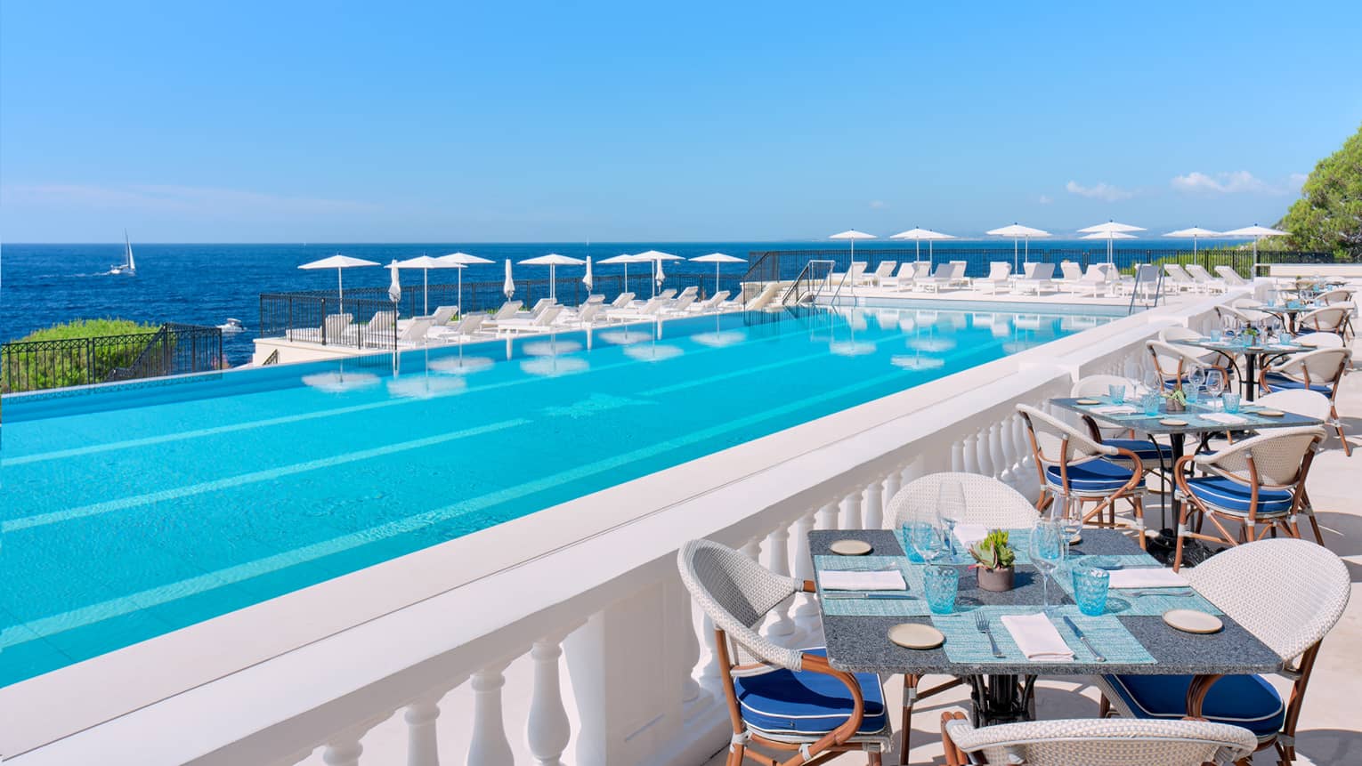 Mediterranean seafront infinity pool surrounded by white tables, lounge chairs, and umbrellas