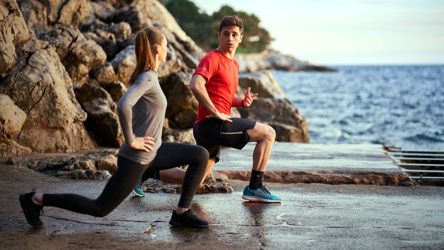 A man trains a woman while doing lunges outside next to the sea and rocks