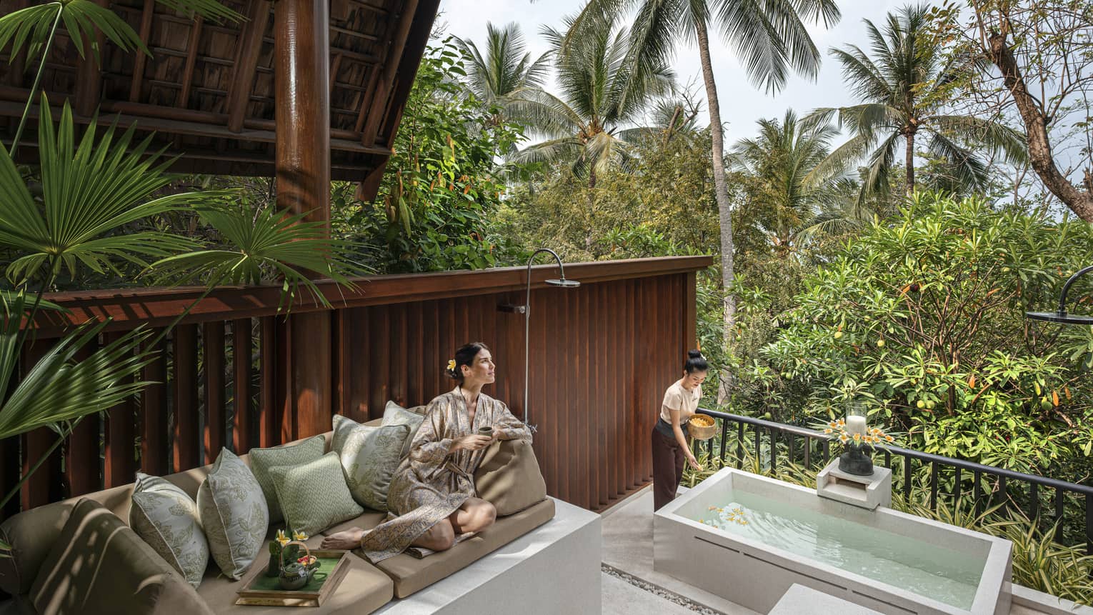 Woman in silk robe lounges on outdoor sofa as spa tech prepares bath