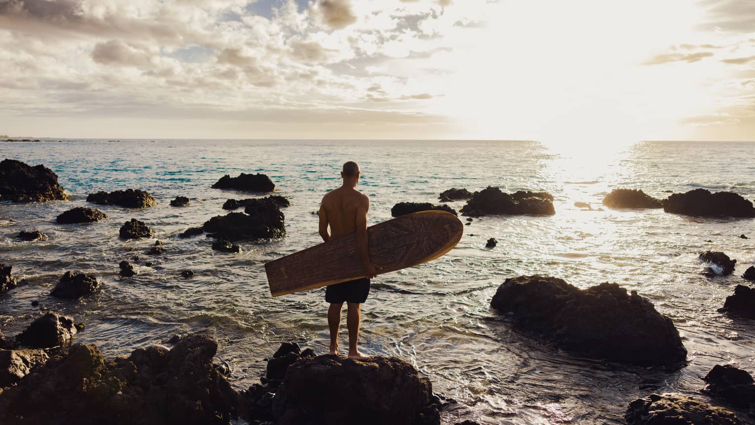Surfboard designer Bonga Perkins stands on craggy beach at sunset with wooden surfboard