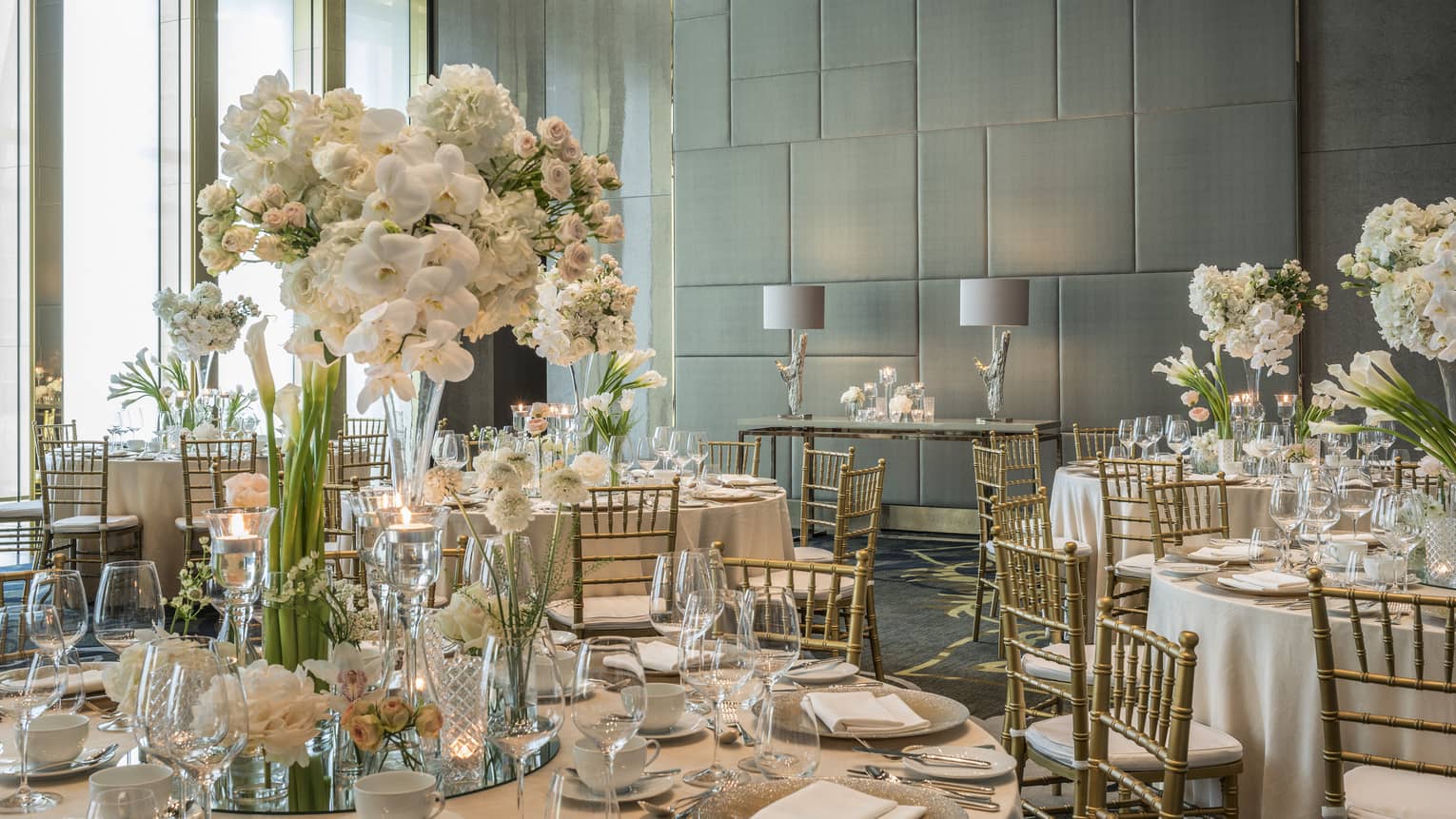 The tables of a wedding reception are set with white tall florals, clear glassware, white plates and silver tableware