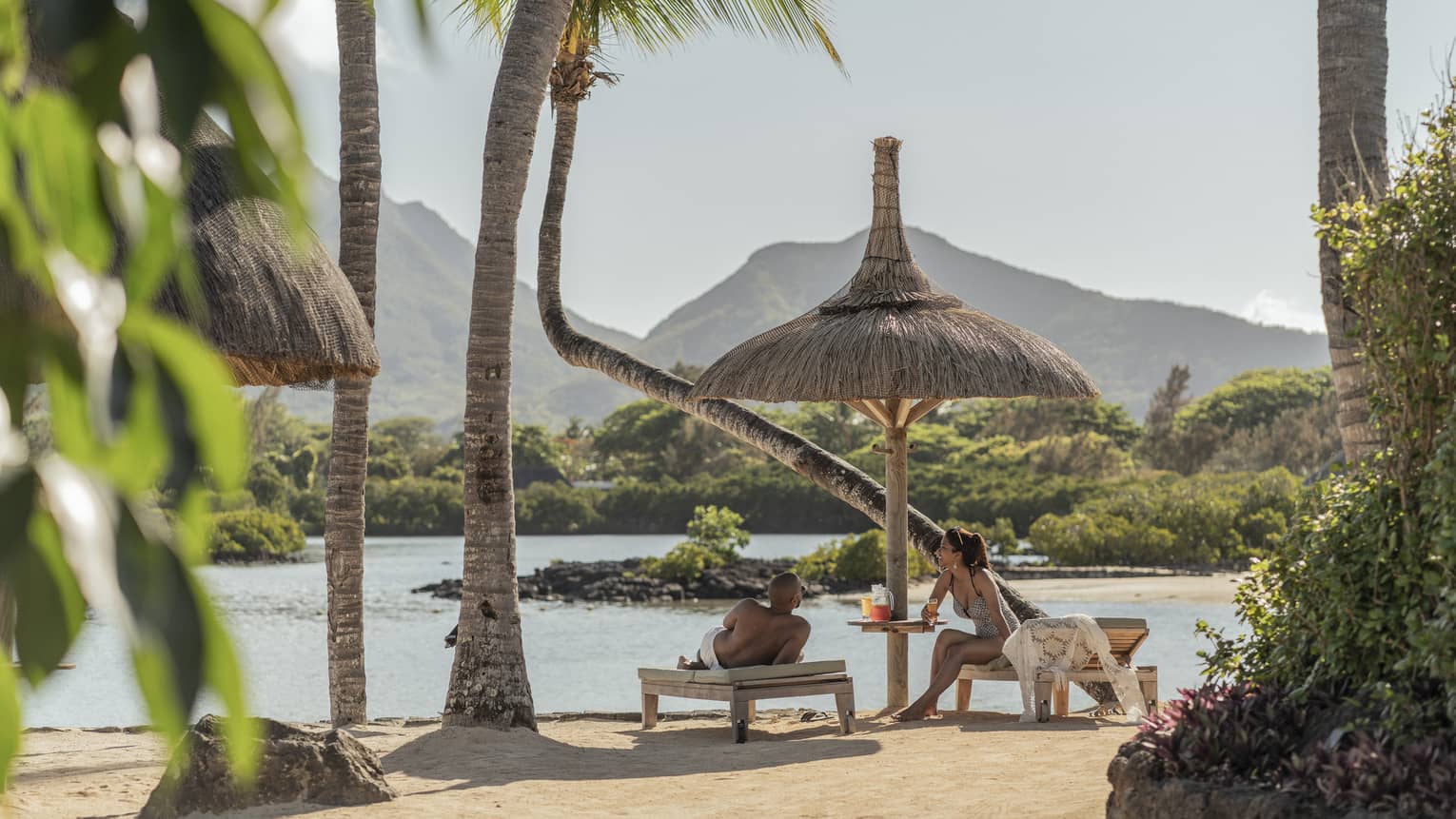 Couple relaxes on oceanfront chaise lounges, mountains and palm trees in the background