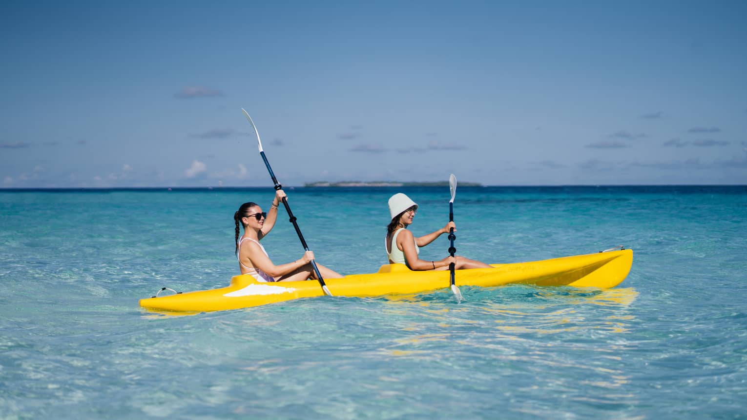 Two people smile as they paddle a yellow kayak in a turquoise lagoon under a clear blue sky; low island in the distance.