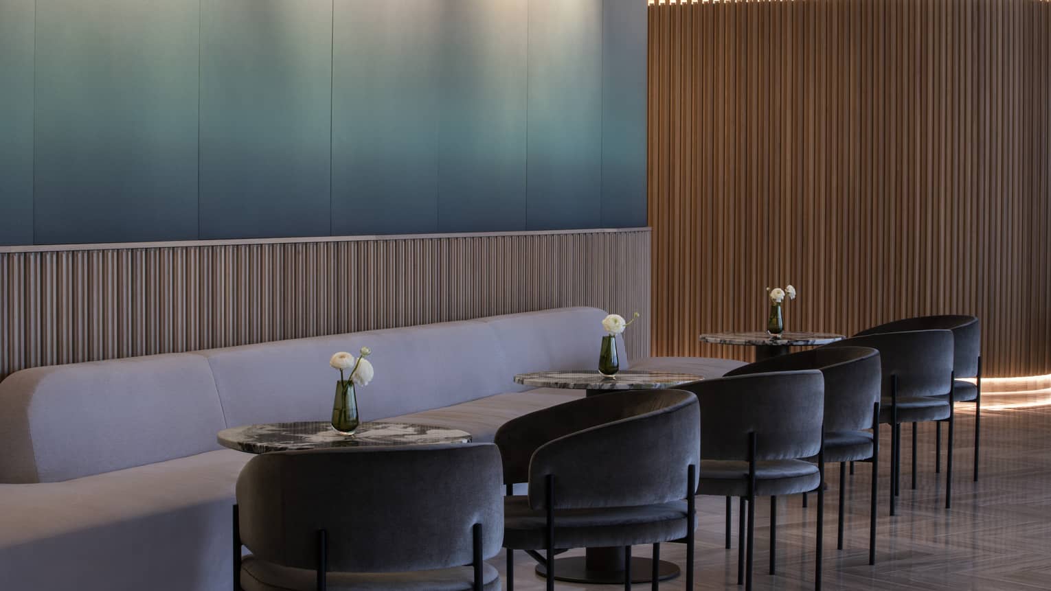Blue-to-white gradient wall, light-grey plush banquet, three round cocktail tables and six dark-grey curved chairs at MARCUS Lounge