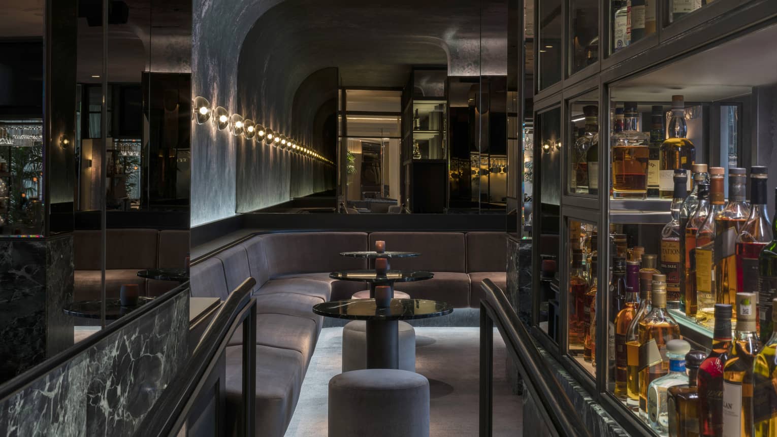 A velvet banquet and three round cocktail tables line the curved wall of MARCUS bar next to glass shelves holding bottle of spirits, all dimly lit