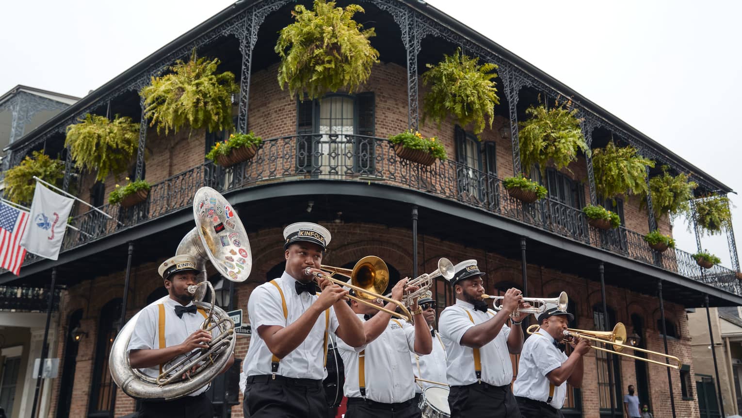 Six-piece brass band plays at the corner of a two-storey building beneath a balcony adorned with flags, ferns and flowers