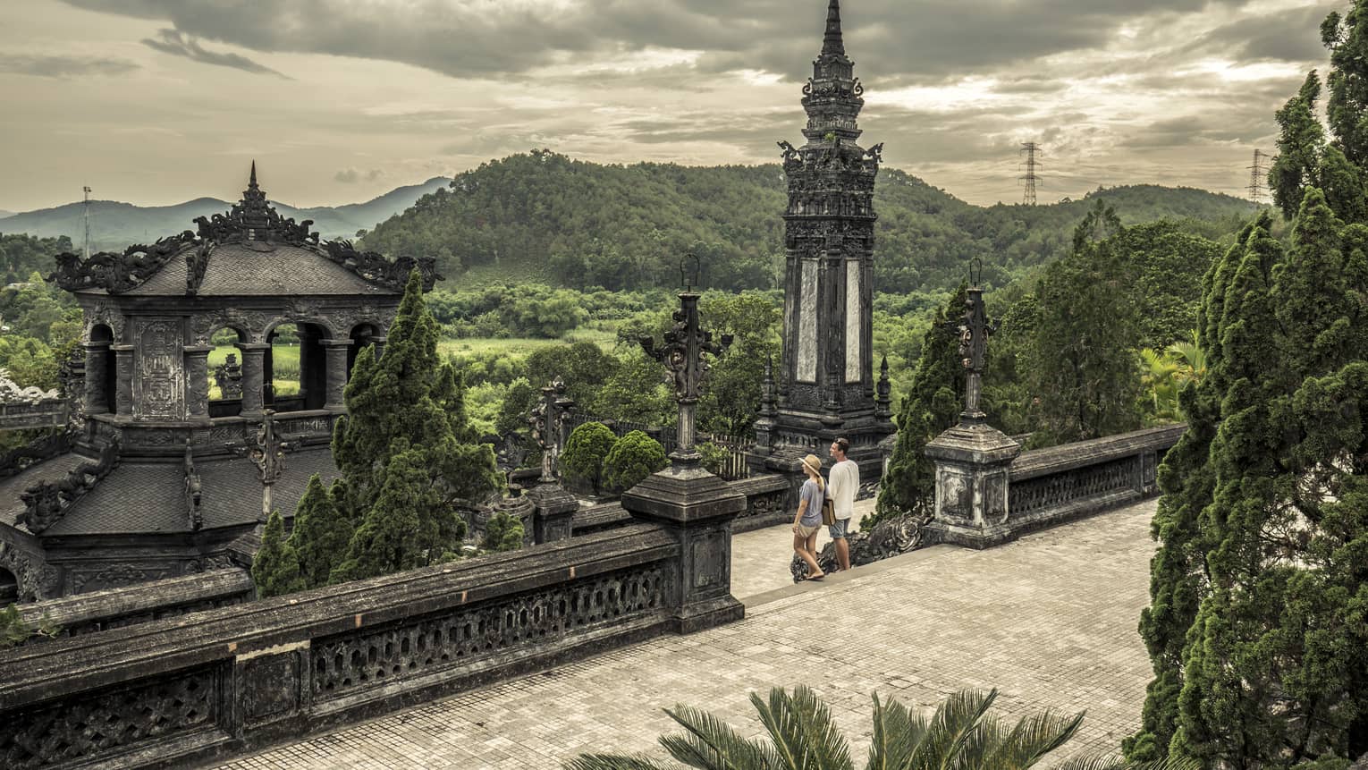 Man and woman walk down steps of historic Imperial City of Hue with stone towers, rolling hills
