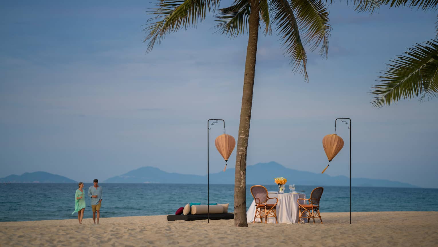 Couple walks on sandy beach by dining table between two palm trees, paper lanterns
