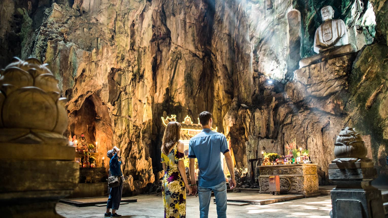 In a cave, three people look up at a large buddha statue atop a shelf in the wall; light streaming in and a shrine below.