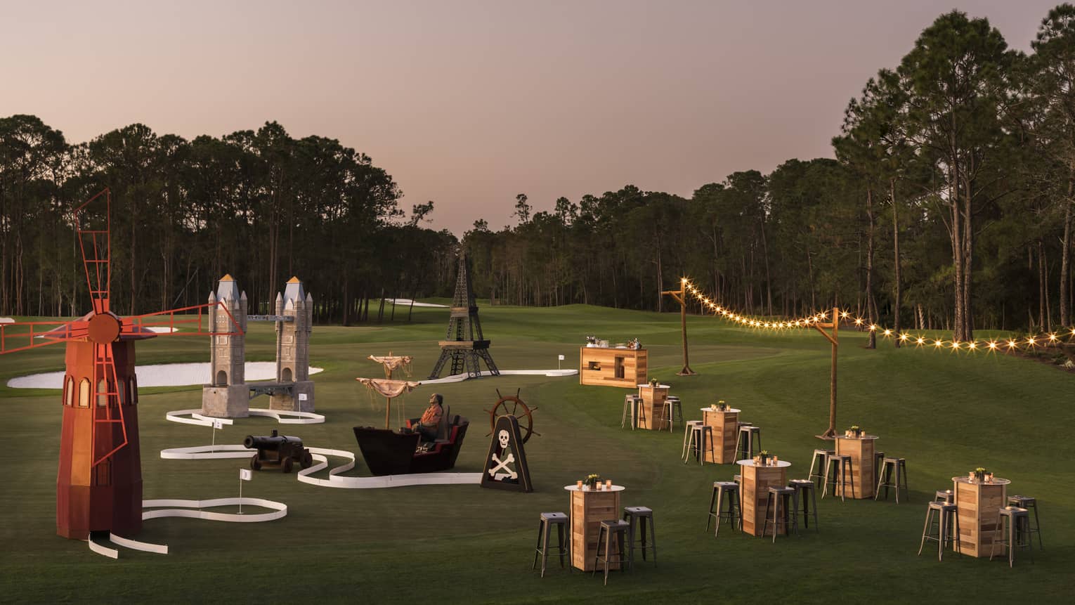 The sun sets over a golf course that's set up with stringed lights and cocktail tables for a putt putt event