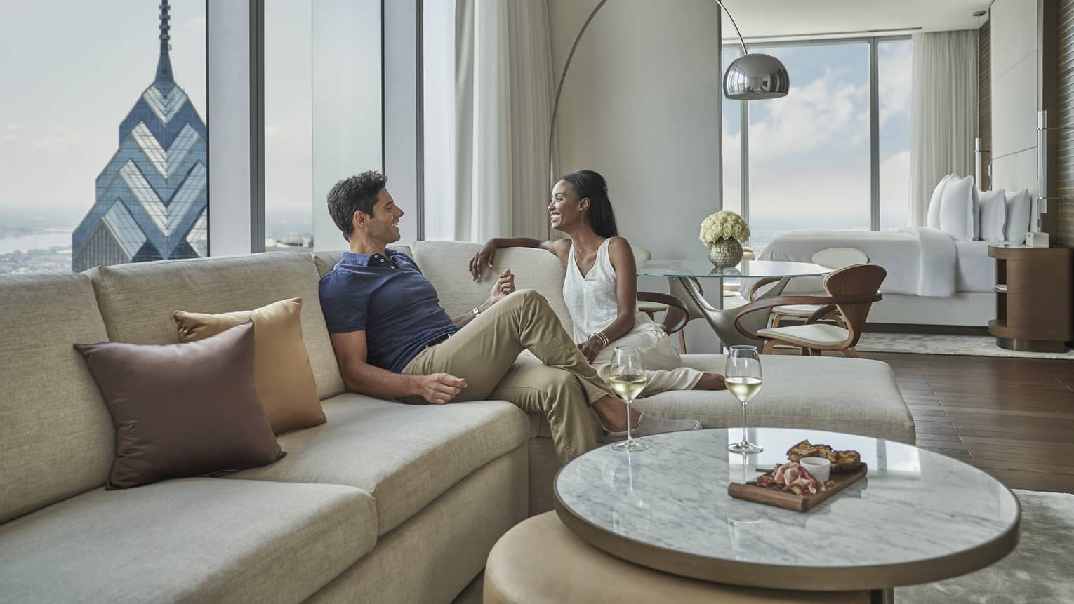 A man and woman lounge on a tan sectional in their modern, light and airy living room adjacent to their bedroom 