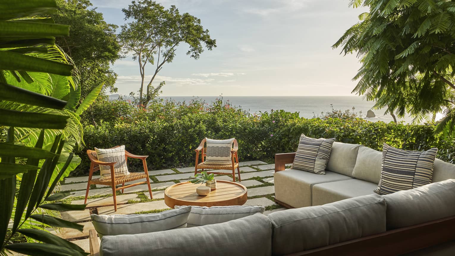Outdoor lounge area with sectional sofa and two chairs, surrounded by green bushes and overlooking the sea