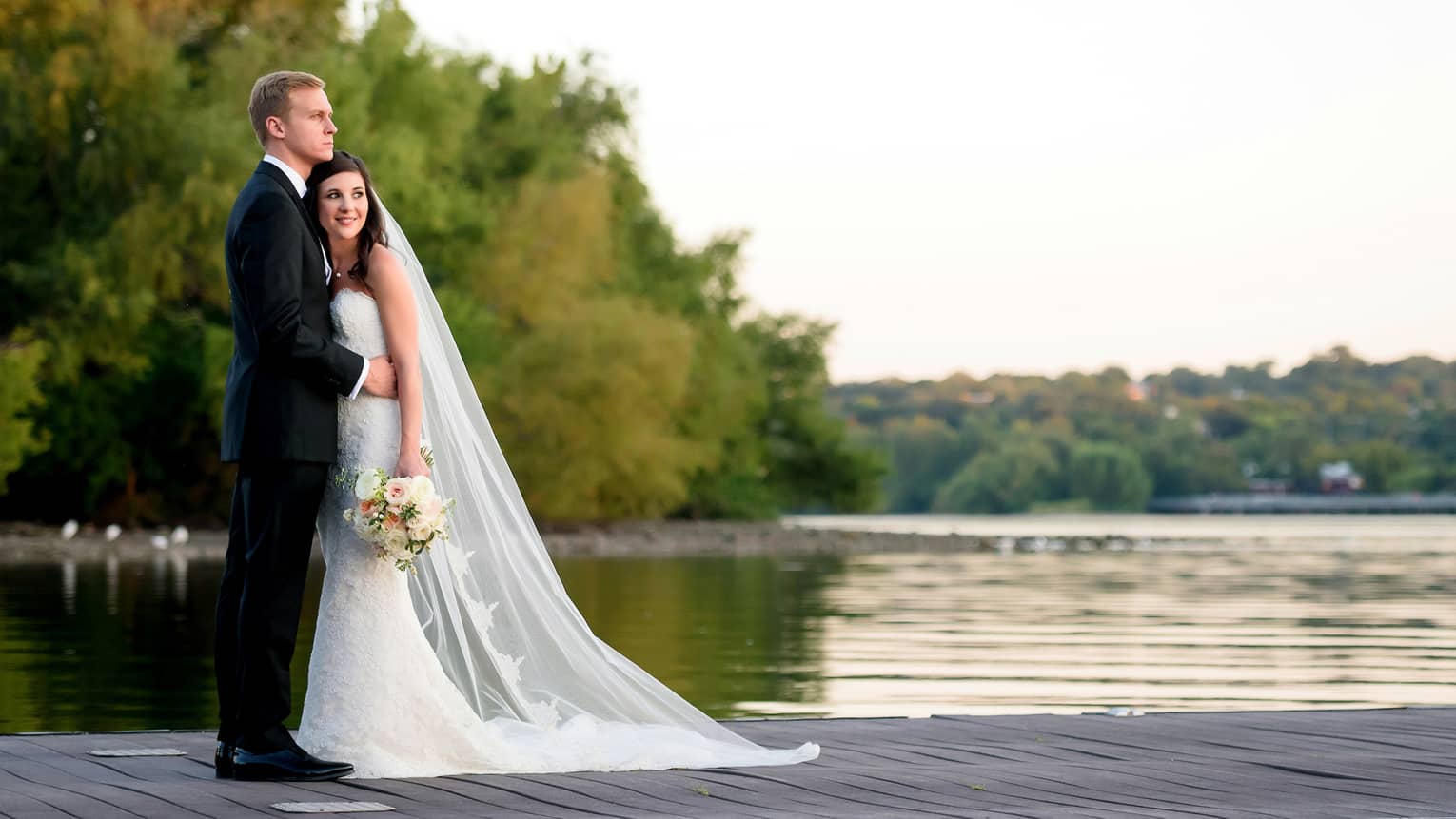Side view of a bride leaning on groom's chest, as they look out over a body of water surrounded by trees