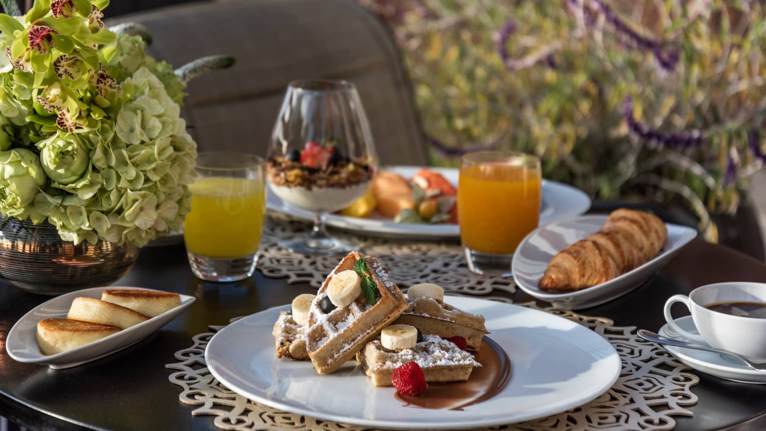 A table filled with breakfast items including sliced waffles topped with fruit, a croissant, two cups of juice, cup of coffee and a glass of yogurt parfait