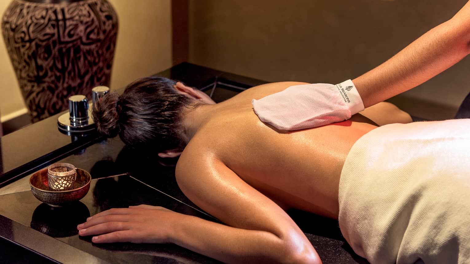 Woman lies face-down on spa table while hand with white mitten massages her back
