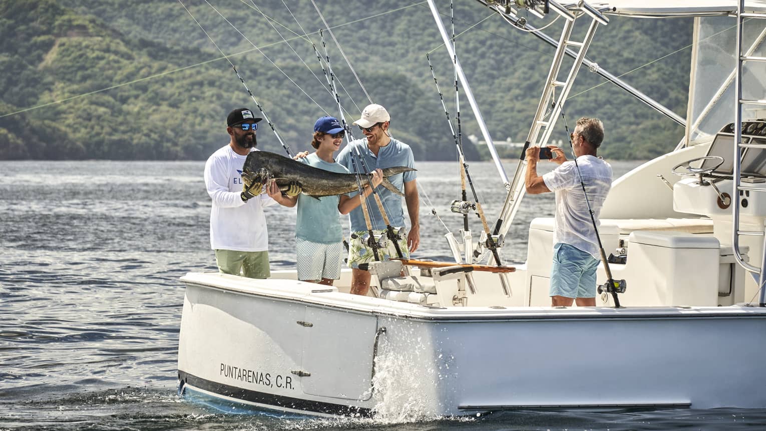 Standing at the back of a fishing boat in the glistening sea against a lush mountain, three smiling people hold a huge fish.