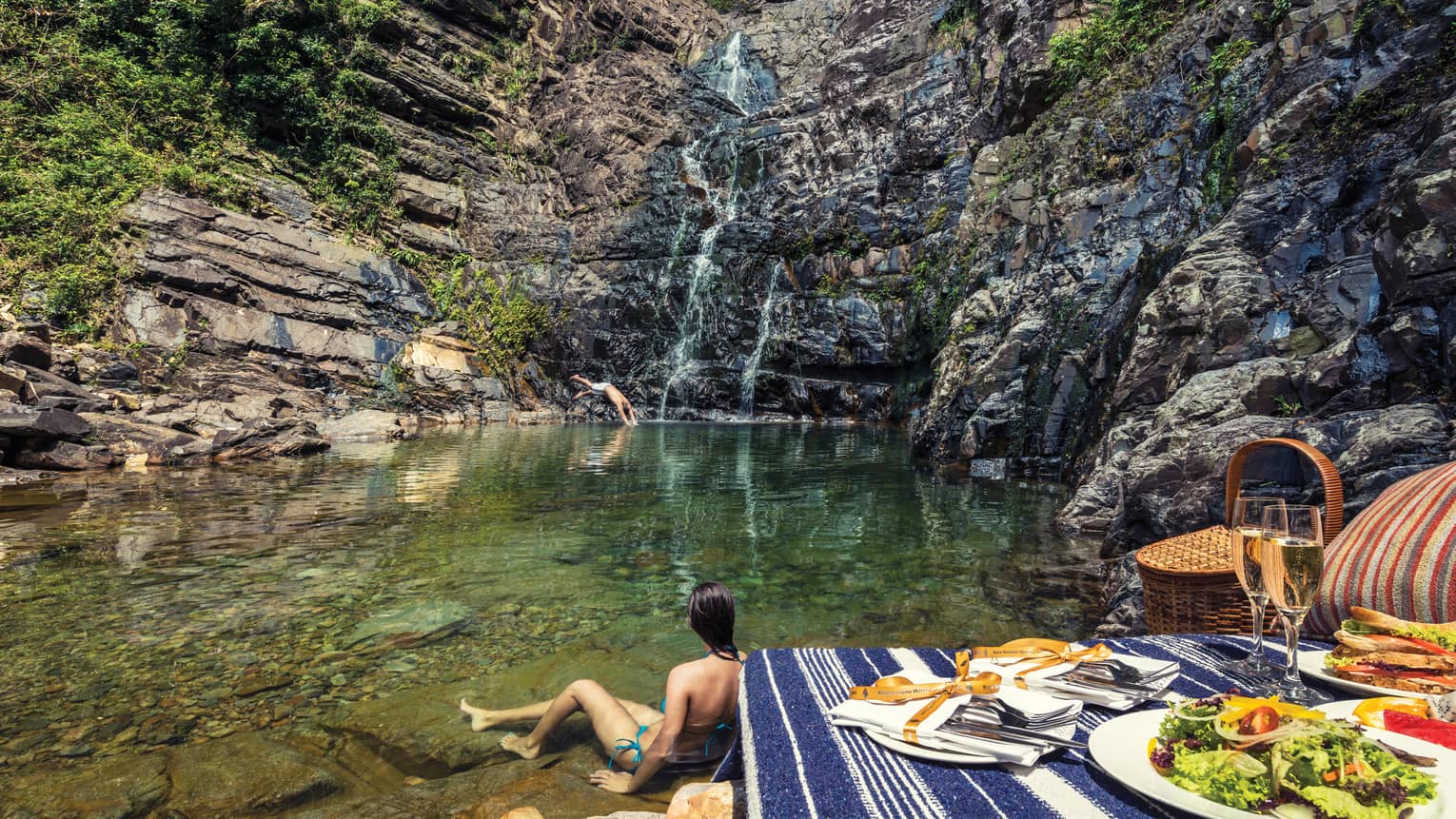 Woman wades in water at bottom of waterfall by picnic lunch set up on rocks